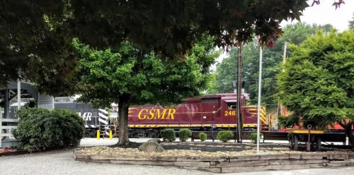 A maroon and yellow train labeled "GSMR" passes by a landscaped area with trees and shrubs.