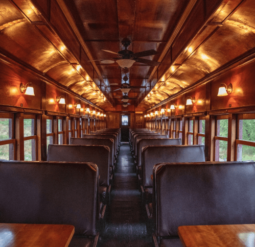 Interior of a vintage train car, featuring wooden paneling, leather seats, and warm lighting along the ceiling.