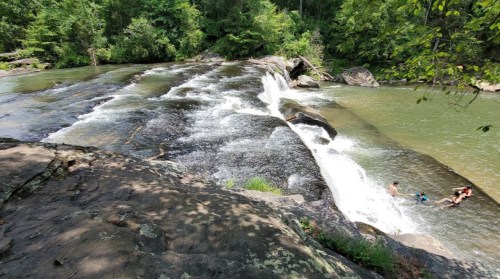 A serene waterfall cascades over smooth rocks into a green river, with two people enjoying the water nearby.