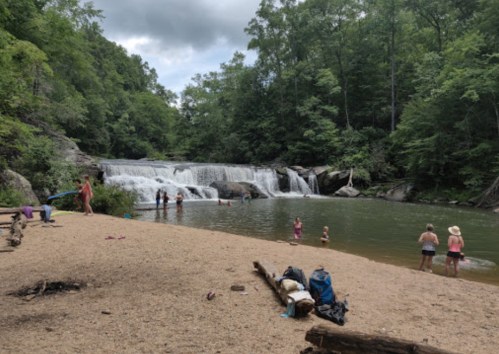 A sandy beach by a waterfall with people swimming and enjoying the water surrounded by lush green trees.