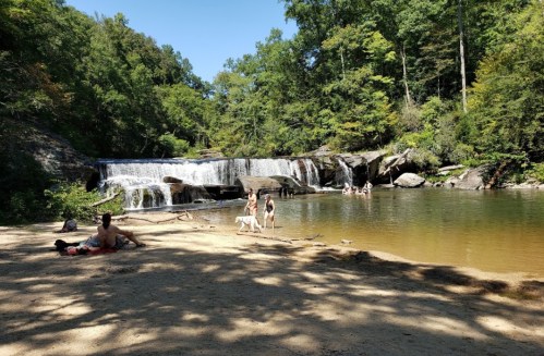 A serene waterfall cascades into a calm pool, with people enjoying the water and sandy shore surrounded by lush greenery.
