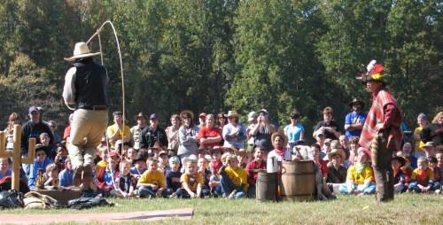 A performer demonstrates lasso skills to a large audience of children and adults in a wooded outdoor setting.