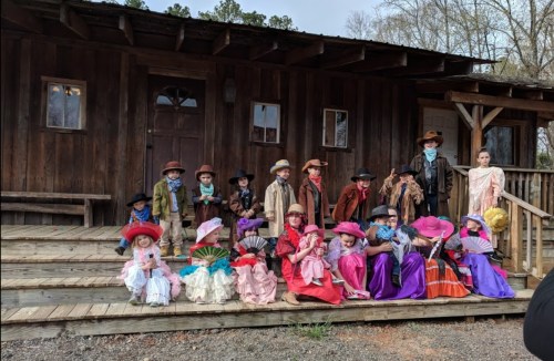 A group of children dressed in colorful costumes posing on the steps of a rustic wooden building.