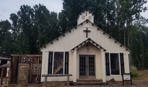 A small white chapel with a cross on top, surrounded by trees, next to a rustic wooden building.