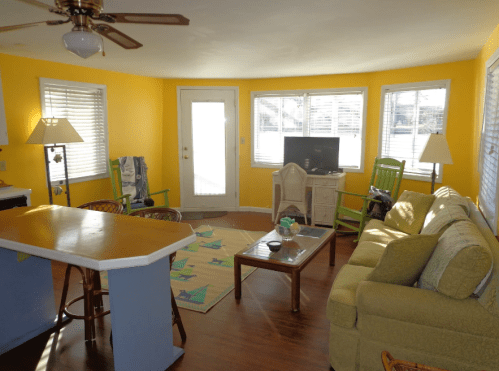 Bright living room with yellow walls, a green sofa, wooden chairs, and large windows letting in natural light.