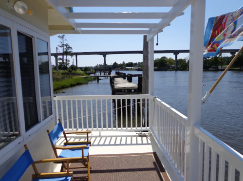 View from a porch overlooking a river, with a dock and bridge in the background under a clear blue sky.