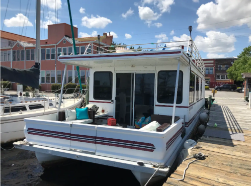 A houseboat docked at a marina, featuring a spacious deck and large windows, surrounded by sailboats and buildings.