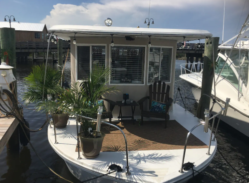 A cozy boat with seating and potted plants, docked in calm waters with a cloudy sky in the background.