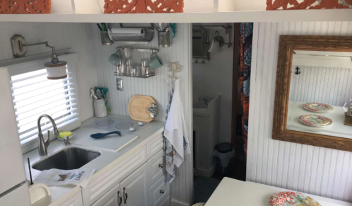 Cozy tiny kitchen with white cabinets, sink, stove, and a small bathroom visible in the background. Bright decor accents.