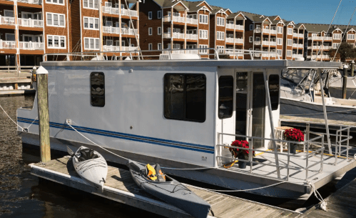 A white houseboat docked at a marina, with kayaks nearby and waterfront apartments in the background.