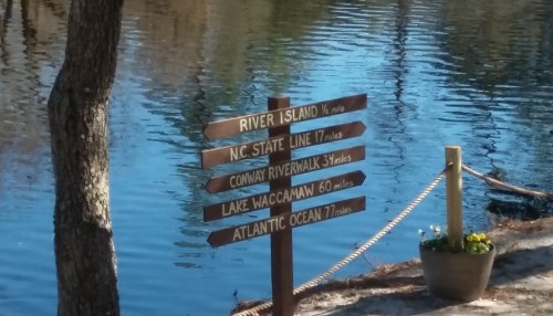 Wooden signpost with directions and distances to River Island, N.C. State Line, Conway Riverwalk, Lake Waccamaw, and Atlantic Ocean.