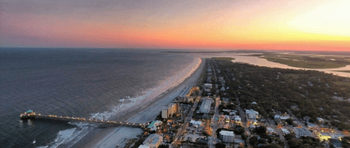 Aerial view of a beach at sunset, with a pier, shoreline, and a coastal town illuminated by evening lights.