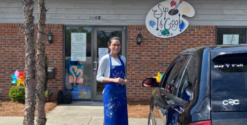 A woman in an apron stands outside a colorful art studio with a sign that reads "Sip & Go." A car is parked nearby.
