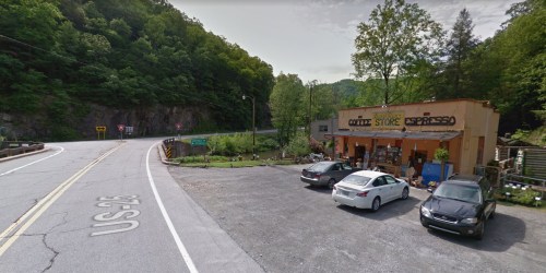 A roadside coffee store with outdoor seating, surrounded by trees and parked cars, along a winding road.