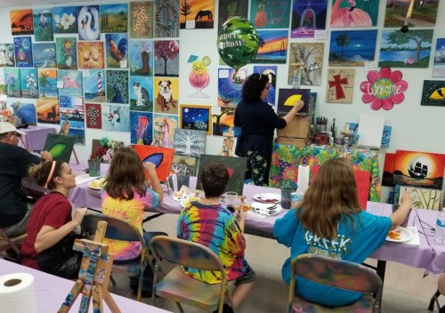 A group of people painting on canvases in a colorful art studio, with various artworks displayed on the walls.