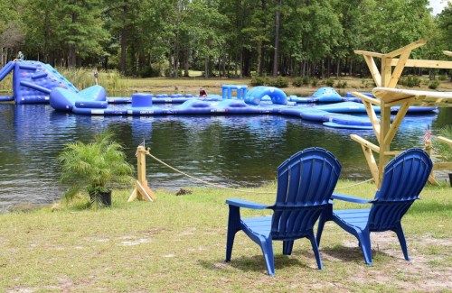 Two blue chairs face a lake with an inflatable water park in the background, surrounded by trees.
