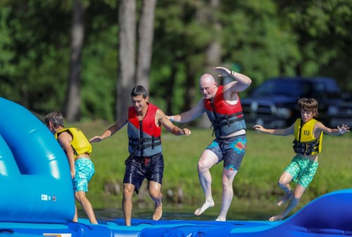 Four people in life jackets play on a water inflatable, enjoying a sunny day by the water.