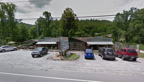 A rustic building with a sign, surrounded by trees and parked cars, along a gravel road in a rural area.
