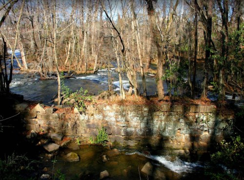 A serene river flows through a wooded area, with bare trees and a stone wall partially visible along the bank.