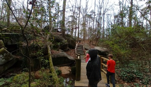 A wooden bridge leads through a wooded area with rocks, as two children explore the nature trail.