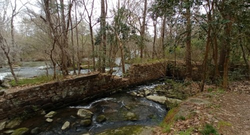 A serene forest scene with a flowing stream, rocky banks, and a stone wall surrounded by bare trees.