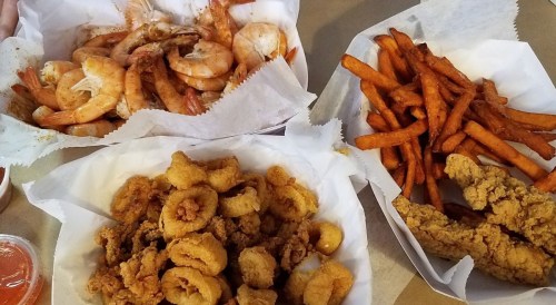 Three trays of seafood: shrimp, fried calamari, and sweet potato fries, served in white paper containers.