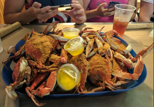 A blue tray filled with seasoned crabs and dipping sauces, with people using phones in the background.