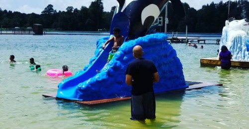 A child slides down a blue whale-shaped water slide into a lake, while others swim nearby.