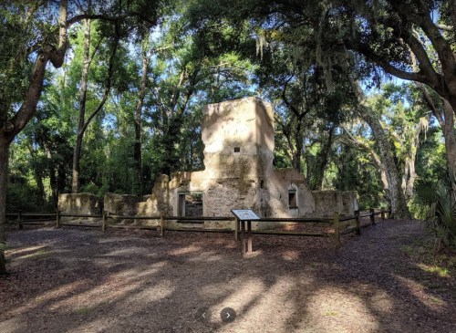Ruins of a stone structure surrounded by lush greenery and trees, with a wooden fence and informational sign nearby.