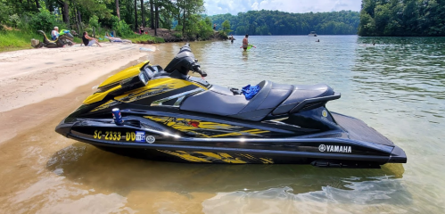 A yellow and black jet ski on a sandy beach by a calm lake, with people swimming and relaxing in the background.