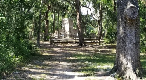 A wooded area with a stone structure and benches surrounded by trees, along a dirt path.