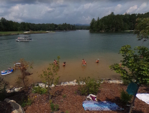 People wade in a calm lake surrounded by trees, with a boat in the distance and towels on the shore.