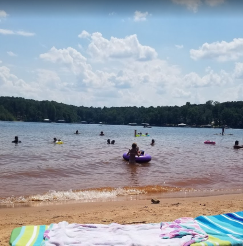 A sandy beach with people swimming in a lake, colorful floats, and a clear blue sky with fluffy clouds.