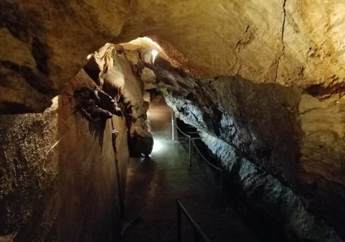 A dimly lit cave with rocky walls and a narrow path leading through water, illuminated by soft light.
