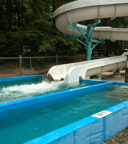 A person slides down a water slide into a blue pool, surrounded by trees and a fence.