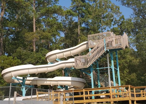 A large water slide with two twisting flumes and a wooden staircase, surrounded by trees and a fenced area.
