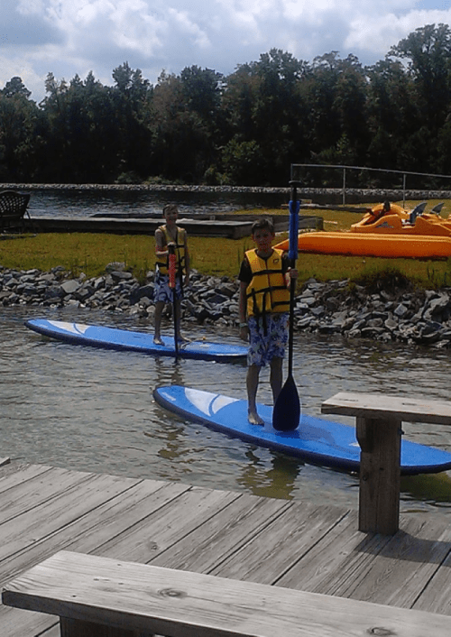 Two children on paddleboards in a calm water area, wearing life jackets, with trees and inflatable boats in the background.