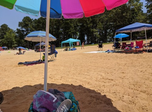 A sandy beach scene with colorful umbrellas, tents, and people relaxing on chairs and towels under a clear sky.