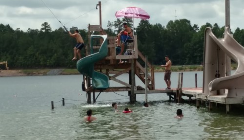 A group of people enjoying a lake with a waterslide, fishing, and swimming on a sunny day.