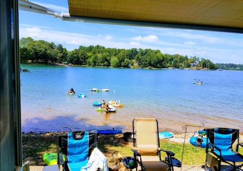 A serene lake view with people on floaties, kayaks, and chairs by the shore under a clear blue sky.