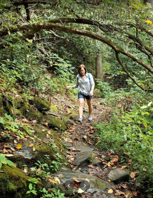 A woman walks along a rocky trail surrounded by lush greenery and fallen leaves in a forested area.