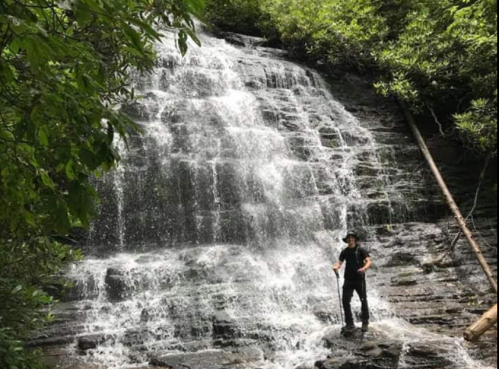 A person stands in front of a cascading waterfall surrounded by lush greenery.