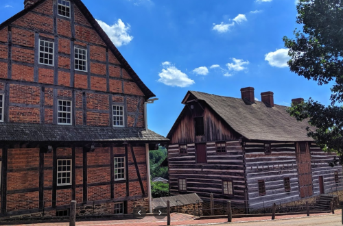 Two historic buildings: one brick with a steep roof, the other wooden with a log cabin style, under a blue sky.
