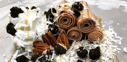 A plate of chocolate rolled pastries topped with whipped cream, pecans, and chocolate pieces, garnished with coconut flakes.