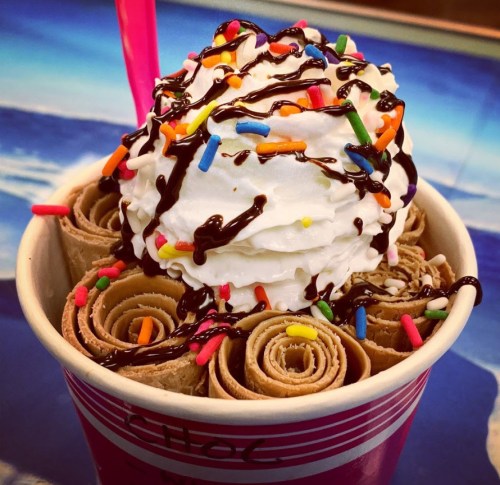 A cup of rolled chocolate ice cream topped with whipped cream, chocolate drizzle, and colorful sprinkles.