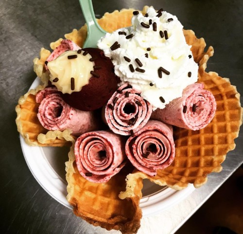 A bowl of rolled strawberry ice cream topped with whipped cream, a chocolate truffle, and chocolate sprinkles in a waffle cup.