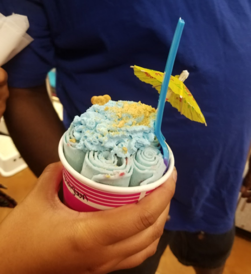 A hand holds a cup of rolled ice cream topped with blue cream, crumbs, a straw, and a small umbrella.