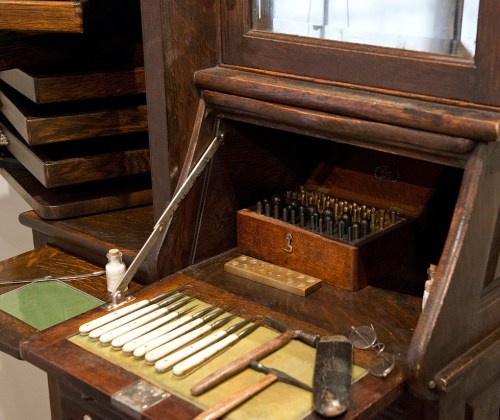 A vintage wooden desk with various tools, a drawer of metal pieces, and a glass display above.