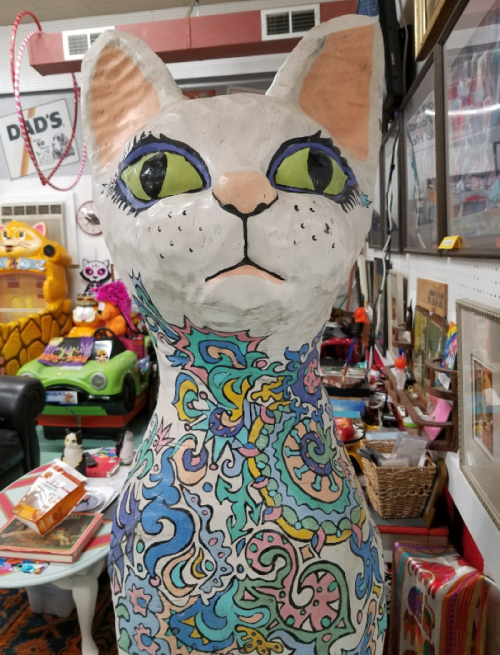 A large, colorful cat sculpture with intricate patterns and bright green eyes, displayed in a quirky shop.