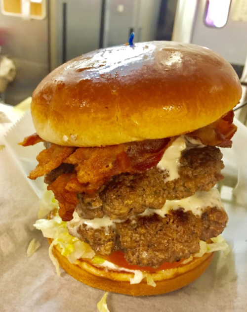 A towering burger with two beef patties, crispy bacon, lettuce, and creamy sauce, all in a shiny bun.